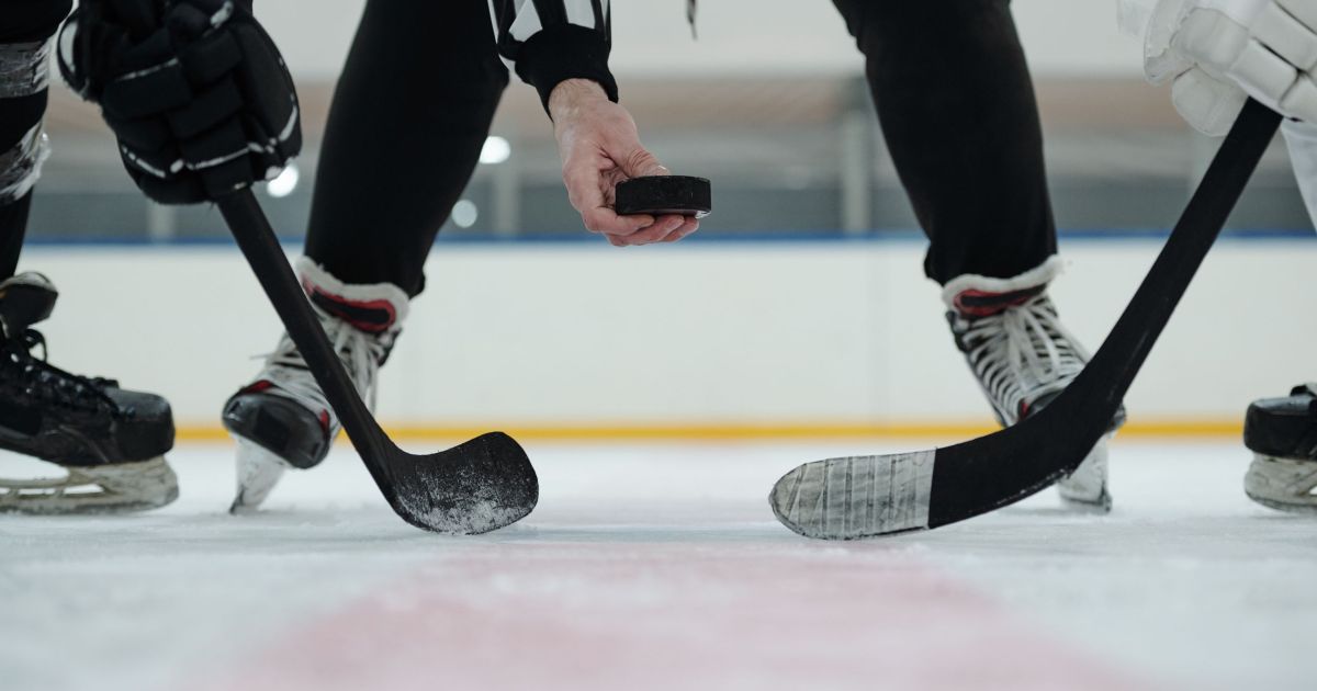 Hand of referee holding puck over ice rink with two players with sticks standing around and waiting for moment to shoot it