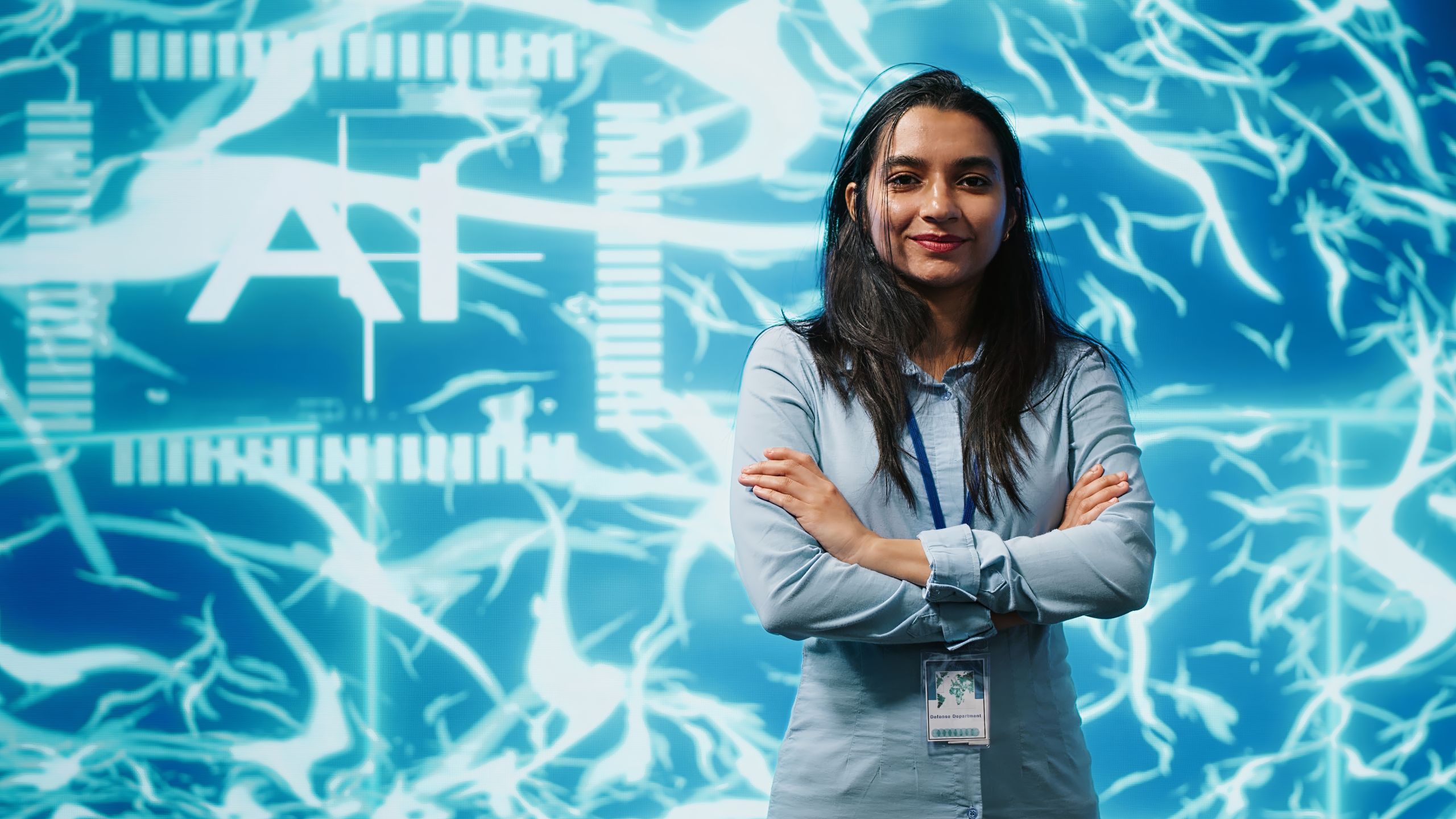 Portrait of smiling IT specialist standing with arms crossed in front of cutting edge display featuring AI and neural network graphics. Researcher in startup doing advancements in AI field, camera A