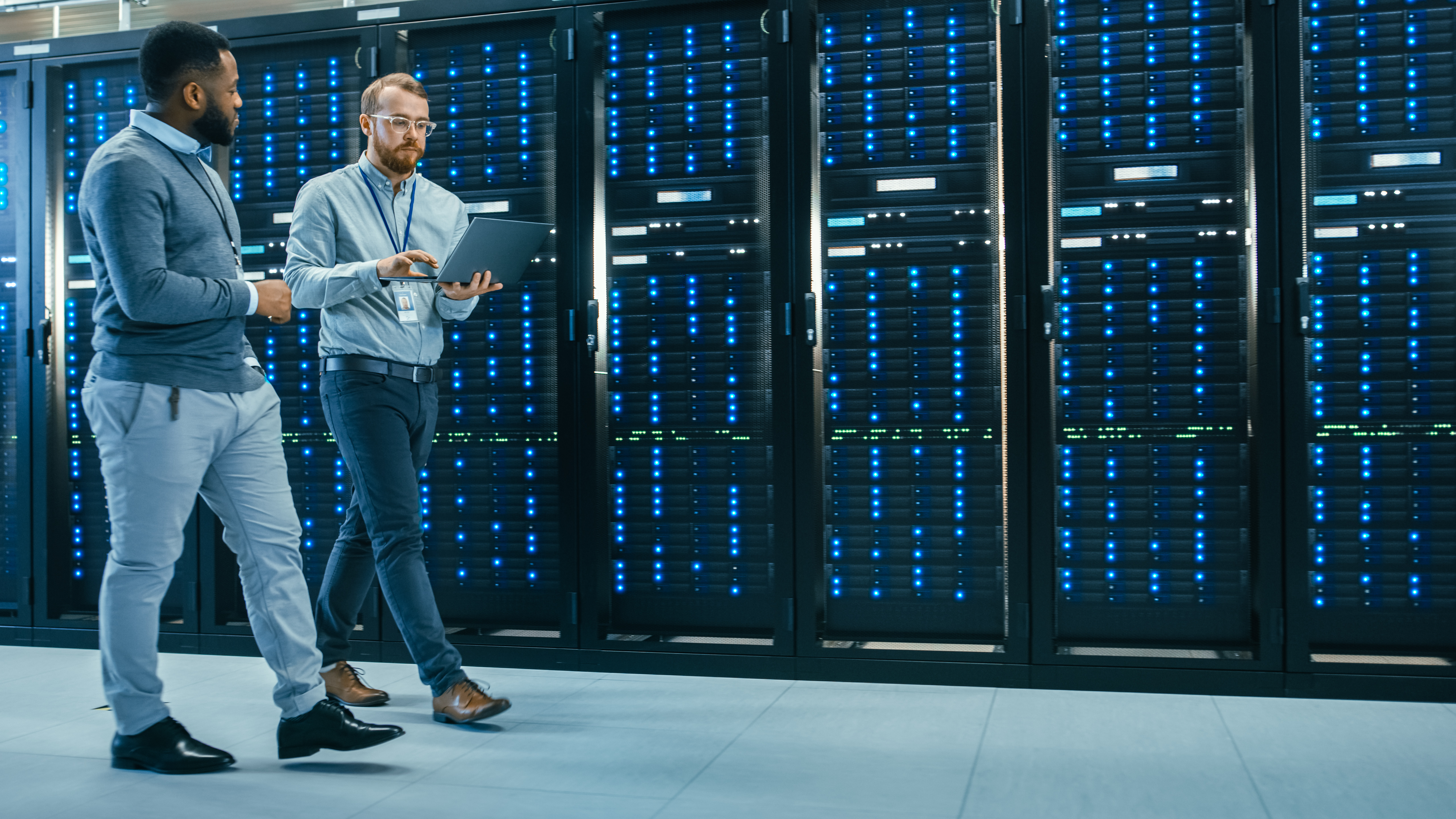 IT Technician with a Laptop Computer and Black Male Engineer Colleague are Talking in Data Center while Walking Next to Server Racks Running Diagnostics or Doing Maintenance Work
